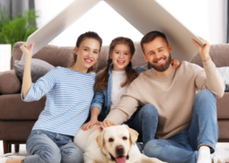 Familie mit Hund steht lachend unter ihrem Hausdach – Symbol für Haus, Wohnen und Tiere