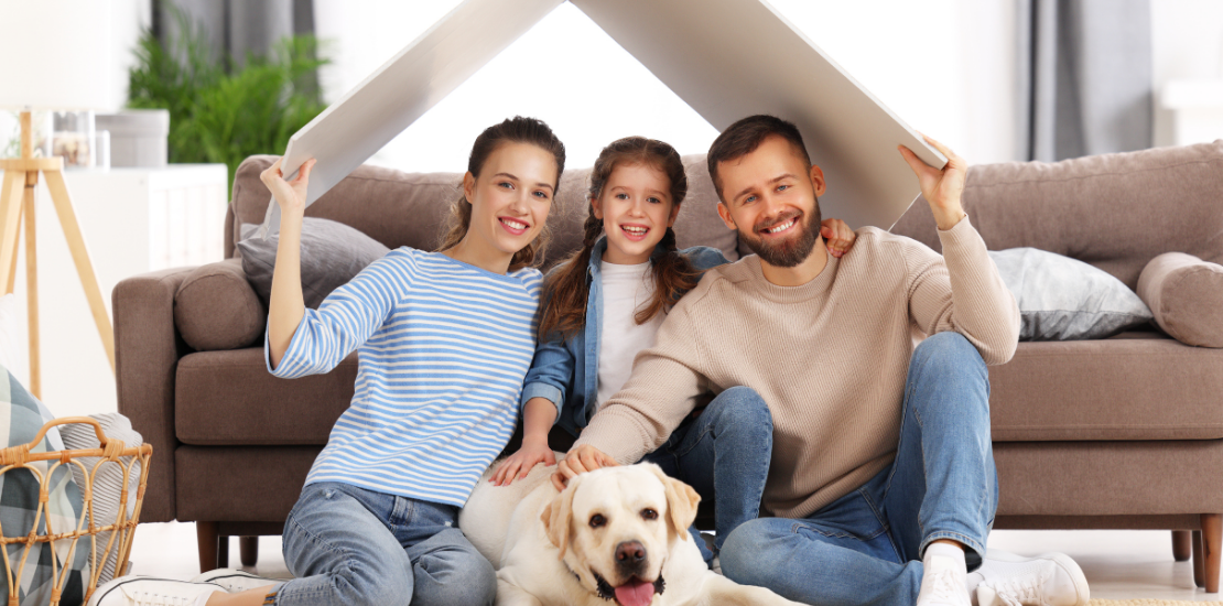 Haus, Wohnen & Tiere 1 Familie mit Hund steht lachend unter ihrem Hausdach – Symbol für Haus, Wohnen und Tiere