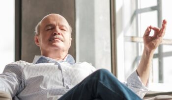 Älterer Mann sitzt entspannt im Sessel und meditiert mit geschlossenen Augen. Symbol für Ruhe, Sicherheit und Gelassenheit im Alltag.