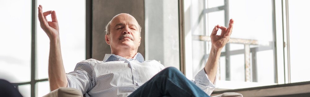 Älterer Mann sitzt entspannt im Sessel und meditiert mit geschlossenen Augen. Symbol für Ruhe, Sicherheit und Gelassenheit im Alltag.