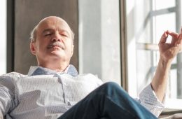 Älterer Mann sitzt entspannt im Sessel und meditiert mit geschlossenen Augen. Symbol für Ruhe, Sicherheit und Gelassenheit im Alltag.